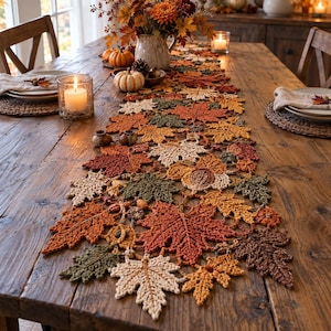 May include: A crochet table runner featuring an intricate pattern of autumn leaves in shades of orange, yellow, brown, cream, and dark green. The textile design incorporates various leaf shapes and small acorn motifs, creating a textured, seasonal display.
