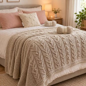 May include: A beige chunky knit throw blanket with a textured cable and bobble pattern draped over a bed. The blanket features vertical braided columns and small raised spheres. Two matching yarn balls sit on the bed surface.