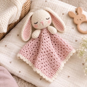 Puede incluir: Una manta de seguridad de ganchillo con una cabeza de conejo color crema, orejas largas, ojos cerrados y mejillas rosadas. La manta adjunta es de color rosa empolvado con un patrón de estrellas texturizado y un borde festoneado crema.