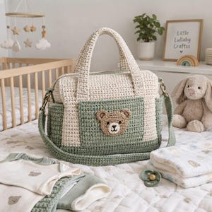 May include: A crochet diaper bag featuring a cream-colored upper section and a sage green lower section with a matching sage green front pocket. The pocket displays a small, brown and cream-colored crocheted teddy bear face appliqué.