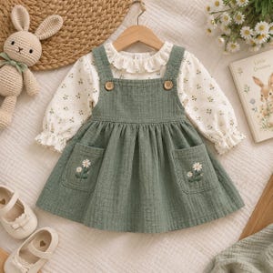 May include: A sage green pinafore dress with two front pockets featuring embroidered white daisies, worn over a long-sleeved cream blouse with a small floral print, a ruffled collar, and ruffled cuffs, hanging on a wooden hanger.