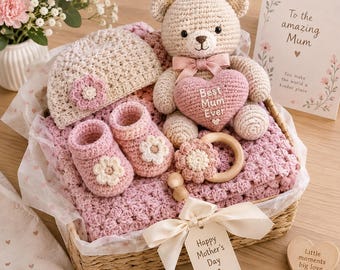 May include: A gift basket containing a crocheted beige bear holding a pink heart with the text Best Mum Ever, a pink crocheted baby blanket, a beige baby hat with a pink flower, pink baby booties with white flowers, and a wooden rattle with a flower.