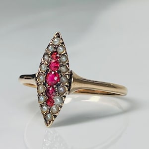 Op de afbeelding: Een gouden ring met een marquise-vormig ontwerp. De ring is versierd met een centrale rij felrode edelstenen, omringd door kleine, glanzende parels. De band en zetting zijn warm goudkleurig, wat een klassieke en elegante uitstraling cre&euml;ert.