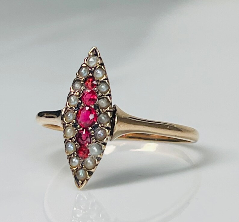 Op de afbeelding: Een gouden ring met een marquise-vormig ontwerp. De ring is versierd met een centrale rij felrode edelstenen, omringd door kleine, glanzende parels. De band en zetting zijn warm goudkleurig, wat een klassieke en elegante uitstraling cre&euml;ert.