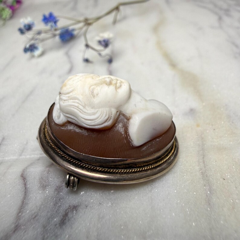 Puede incluir: Broche de camafeo ovalado con una figura blanca tallada sobre un fondo marr&oacute;n, engastada en un marco plateado con un borde de cuerda dorada. El broche tiene un cierre de alfiler en la parte posterior.