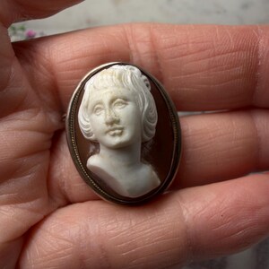 Puede incluir: Broche de camafeo ovalado con un retrato en relieve blanco de una mujer con pelo corto, sobre un fondo marr&oacute;n. El camafeo est&aacute; enmarcado por un borde de metal plateado. Una joya cl&aacute;sica.