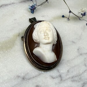 Puede incluir: Colgante de camafeo ovalado con un busto blanco tallado de una mujer sobre un fondo marr&oacute;n. El colgante tiene un marco plateado con un borde decorativo y una argolla para una cadena. Una joya cl&aacute;sica.