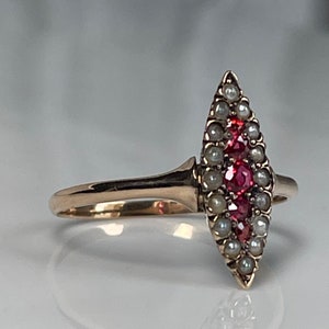 Op de afbeelding: Een gouden ring met een marquisevormige cluster van kleine parels en helderrode edelstenen. Het ontwerp van de ring is langwerpig, met de edelstenen en parels symmetrisch gerangschikt. De band is van eenvoudig, gepolijst goud.