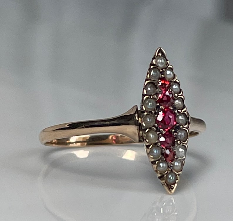 Op de afbeelding: Een gouden ring met een marquisevormige cluster van kleine parels en helderrode edelstenen. Het ontwerp van de ring is langwerpig, met de edelstenen en parels symmetrisch gerangschikt. De band is van eenvoudig, gepolijst goud.