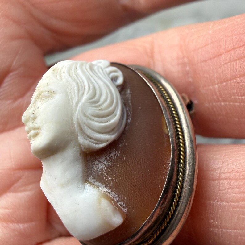Puede incluir: Broche de camafeo vintage con un perfil tallado de una mujer con cabello suelto. El camafeo est&aacute; engastado en un marco plateado con un borde de cuerda dorada. El perfil de la mujer es blanco sobre un fondo marr&oacute;n.