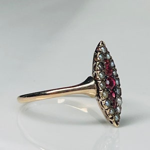 Op de afbeelding: Een gouden ring met een marquise-vormige voorkant. De voorkant is versierd met kleine, ronde parels en levendige rode edelstenen. De ringband is slank en gepolijst, waardoor het licht wordt gereflecteerd.