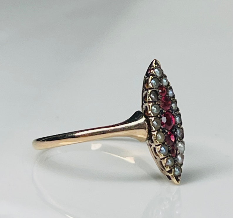 Op de afbeelding: Een gouden ring met een marquise-vormige voorkant. De voorkant is versierd met kleine, ronde parels en levendige rode edelstenen. De ringband is slank en gepolijst, waardoor het licht wordt gereflecteerd.