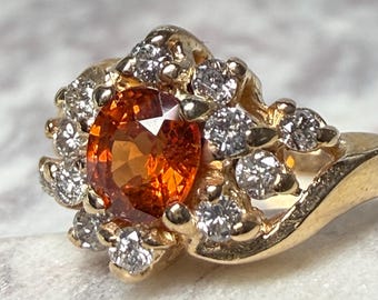 May include: A gold ring featuring an oval, orange gemstone surrounded by small, round, clear gemstones. The ring's band is a simple, polished gold. The gemstones are set in a floral-like arrangement.