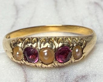 May include: A gold ring featuring a row of five gemstones. The ring alternates between a light-colored pearl and a deep red gemstone. The band has an ornate design along the top. The ring is set against a marble background.