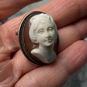 Puede incluir: Broche de camafeo ovalado con un busto femenino blanco sobre un fondo marr&oacute;n. El camafeo est&aacute; enmarcado por un borde de metal dorado con un borde decorativo en forma de cuerda. El broche tiene un cierre de alfiler en la parte posterior.