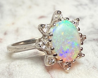 May include: A silver ring featuring a large, oval opal gemstone with iridescent colors. The opal is surrounded by a halo of smaller diamonds, creating a starburst design. The ring is set on a white marble surface.