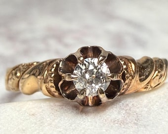May include: A gold ring with a central diamond. The ring features a floral-shaped setting around the diamond and a twisted band with engraved details. The ring is set on a white surface.