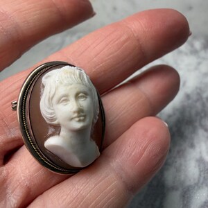 Puede incluir: Broche de camafeo ovalado con un retrato en relieve blanco de una mujer. El camafeo est&aacute; engastado sobre un fondo marr&oacute;n dentro de un marco dorado, con un cierre de alfiler en la parte posterior. Una joya cl&aacute;sica.