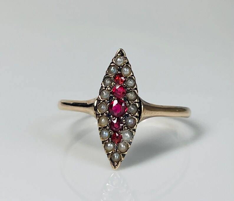 Op de afbeelding: Een gouden ring met een marquise-vormig middelpunt. Het is versierd met kleine, ronde parels en levendige rode edelstenen. De ring heeft een eenvoudige, gepolijste gouden band.