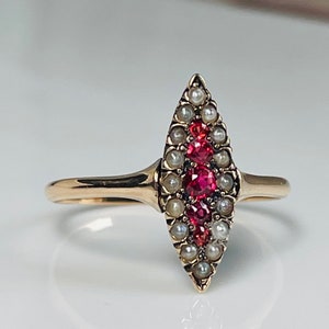 Op de afbeelding: Een gouden ring met een marquise-vormig ontwerp. De ring is versierd met een centrale rij felrode edelstenen, omrand door kleine, glanzende parels. De band is effen, gepolijst goud.