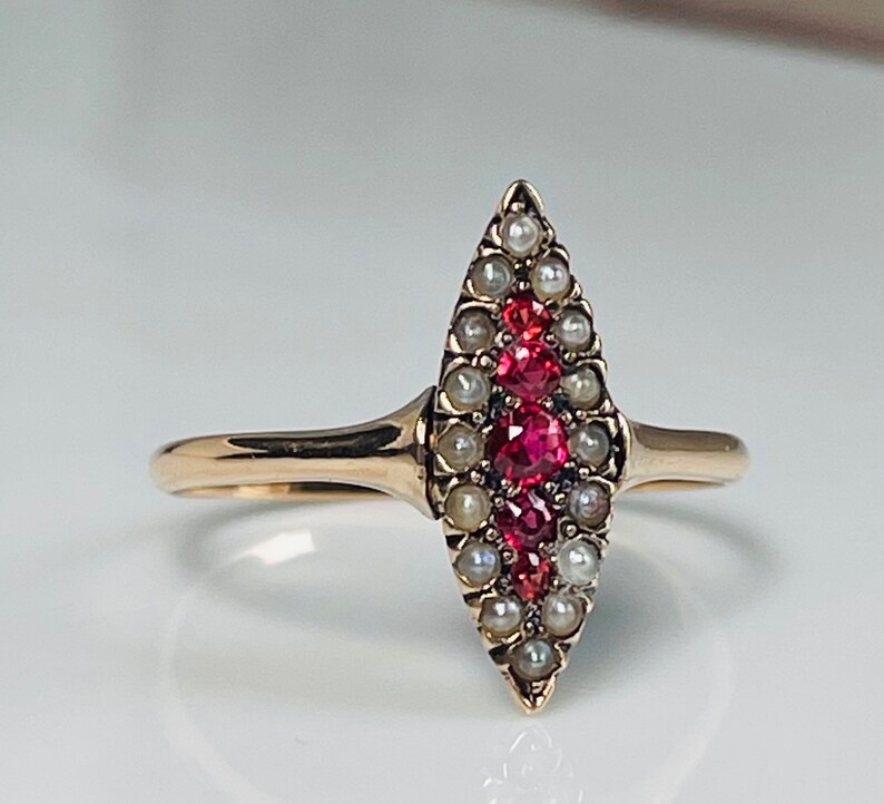 Op de afbeelding: Een gouden ring met een marquise-vormig ontwerp. De ring is versierd met een centrale rij felrode edelstenen, omrand door kleine, glanzende parels. De band is effen, gepolijst goud.