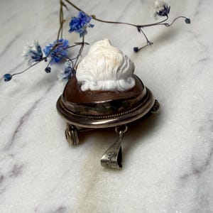 Puede incluir: Colgante camafeo vintage con una figura de concha blanca tallada sobre una base marr&oacute;n, enmarcada en metal plateado. El colgante tiene un peque&ntilde;o lazo y una anilla para una cadena. Flores azules en el fondo.