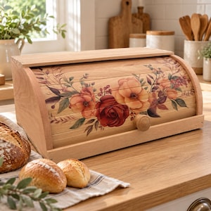 May include: A light-colored wooden bread box with a roll-top door featuring a printed floral design of red and orange roses with green leaves. The box has a small round wooden knob on the front and sits on a wooden surface near fresh bread.