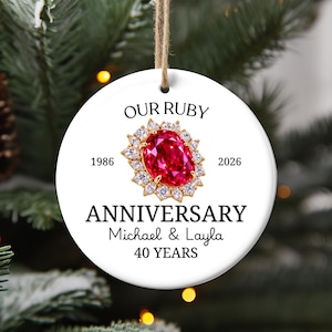Op de afbeelding: Een rond wit keramisch ornament met een centrale afbeelding van een ovale robijn omringd door heldere diamanten. De tekst luidt OUR RUBY ANNIVERSARY, 1986, 2026, Michael & Layla en 40 YEARS. Het ornament hangt aan een bruin touwtje.