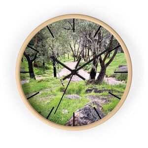 May include: A round wall clock with a light wood frame featuring a printed image of a forest landscape with green grass, dark tree trunks, and a dirt path. The clock has thin black hour, minute, and second hands and black tick marks.