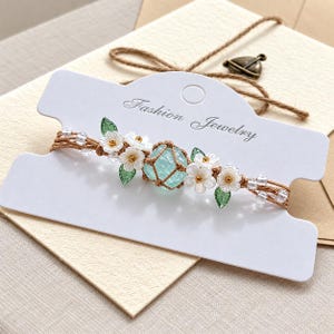 Può includere: Un braccialetto su un cartoncino bianco con la scritta Fashion Jewelry. Il braccialetto presenta una perla centrale azzurra testurizzata in corda marrone, affiancata da fiori bianchi con centri dorati e piccole foglie verdi, con perline trasparenti.