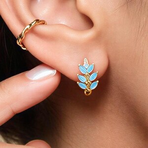 May include: A gold-toned ear cuff with a twisted texture and a gold-toned stud earring featuring a vertical arrangement of six light blue enamel leaves. The stud earring has a small gold loop at the bottom, and both pieces are worn on the ear.