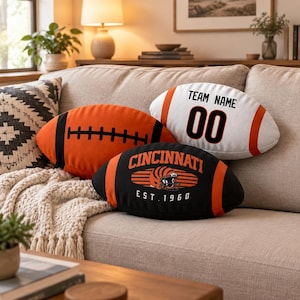 Op de afbeelding: Drie football-vormige sierkussens. Eén is oranje met zwarte veters, één is wit met oranje strepen en de tekst "TEAM NAME 00", en één is zwart met oranje strepen, een helm-afbeelding en de tekst "CINCINNATI EST. 1960".