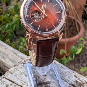 May include: A wristwatch with a rose gold-tone case and a dark brown leather strap. The dial features a textured, gradient copper-to-black finish, rose gold-tone hands and indices, and an open-heart aperture revealing the mechanical movement.
