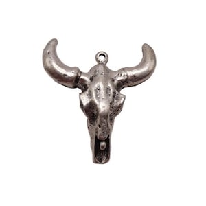 May include: A silver-colored bull skull pendant with curved horns and a textured surface. The pendant has a loop at the top for attaching to a chain or cord. The skull is detailed with eye sockets and a nasal cavity.