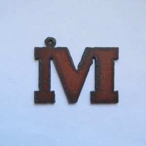May include: A rusty metal letter M with a hole at the top for hanging. The letter is a dark brown color with a slightly orange hue.