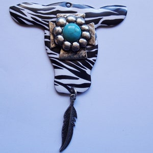 May include: A black and white zebra patterned metal cow skull with a turquoise stone and silver embellishment. A silver feather dangles from the bottom.