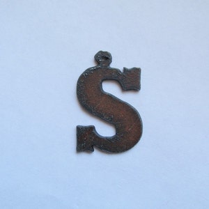 May include: A rusty metal letter S with a decorative design. The letter is a dark brown color and has a rough texture.