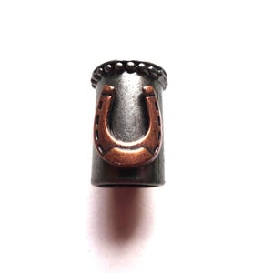 May include: A silver metal toggle with a copper horseshoe charm. The toggle has a beaded edge.