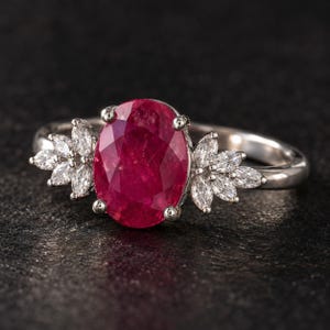 May include: A silver-toned ring featuring a large, oval-cut, deep red gemstone at the centre. The ring is accented on both sides by a cluster of three marquise-cut, clear white stones set in a floral-like arrangement on a polished metal band.