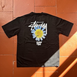 Peut inclure: Un t-shirt noir à manches courtes avec un motif au dos. Le design comprend le logo Stussy blanc au-dessus d'une fleur de marguerite bleue et jaune, avec le texte "Increase the Peace" imprimé en lettres blanches sous le motif floral.