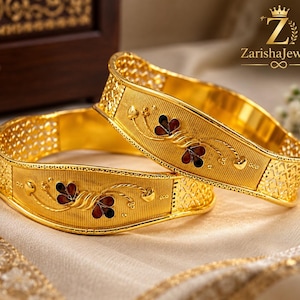 Puede incluir: Dos brazaletes de oro con una banda central texturizada y un motivo floral en esmalte rojo y azul oscuro. Los lados de los brazaletes tienen un patrón de celosía calado. El texto ZarishaJewels con un icono de corona es visible arriba.
