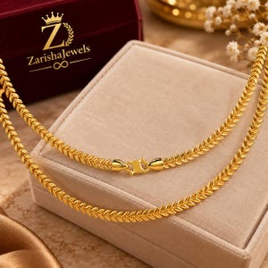 Puede incluir: Un collar de cadena dorada con un patrón repetitivo de hojas y un cierre pulido. El collar se muestra con un logotipo que dice ZarishaJewels con un icono de corona y un símbolo de infinito sobre un joyero de terciopelo rojo oscuro.
