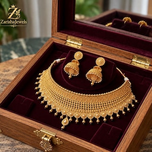 Puede incluir: Un collar de oro y un par de pendientes jhumka a juego con filigrana y abalorios, presentados en una caja de madera con forro de terciopelo rojo oscuro. La tapa de la caja muestra el logotipo de ZarishaJewels en letras doradas.