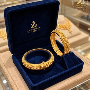 Puede incluir: Dos brazaletes de oro con superficie texturizada de cuentas presentados en un joyero de terciopelo azul marino. El interior de la tapa muestra el texto dorado ZarishaJewels con un logotipo de corona e infinito. Los brazaletes tienen un patrón de puntos.