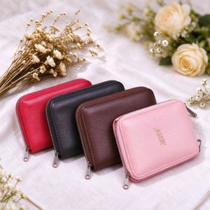 May include: Four rectangular zippered wallets with a textured finish, arranged in a row. The colors are red, black, dark brown, and light pink. The light pink wallet features the gold-colored text JANET embossed on the front surface.