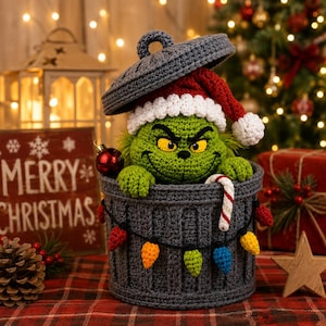 May include: A crocheted green creature with yellow eyes and a wide grin, wearing a red and white Santa hat, sits inside a dark gray crocheted trash can. The can is decorated with a string of colorful crocheted Christmas lights and a candy cane.