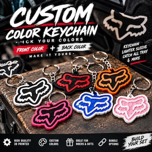 May include: A collection of custom 3D printed keychains featuring the Fox Racing logo in various color combinations including black and white, black and red, black and orange, black and pink, blue and white, and pink and white on a wooden surface.