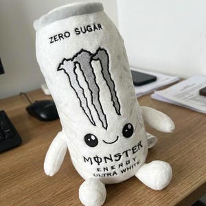 May include: A white plush toy shaped like a Monster Energy Ultra White can. It features a grey top, the Monster logo, and the text "ZERO SUGAR" and "MONSTER ENERGY ULTRA WHITE" printed on the front. The plush has two small arms, two legs, and a face with two black eyes and a smiling mouth.