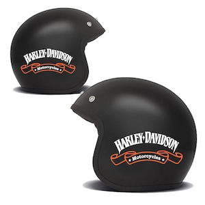 Może przedstawiać: Dwa matowe czarne otwarte kaski motocyklowe z białym logo Harley-Davidson nad pomarańczowym banerem z napisem Motorcycles w kolorze białym. Każdy kask posiada srebrny metalowy zatrzask w okolicy skroni przeznaczony na akcesoria.