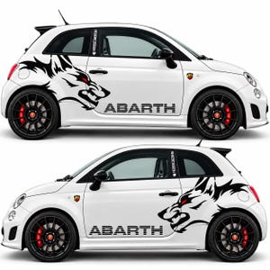 Peut inclure: Une voiture à hayon Abarth blanche arborant un grand motif noir représentant une tête de loup menaçante aux yeux rouges sur l'aile arrière, avec le mot ABARTH en lettres noires sur la porte, dotée de jantes noires à rayons et d'étriers rouges.